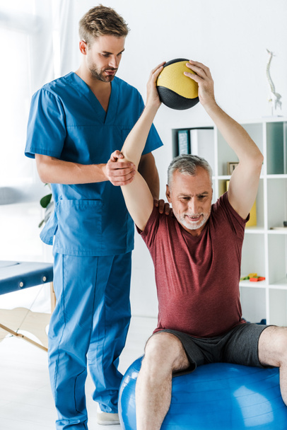 beau médecin debout près d'un patient mature faisant de l'exercice sur une balle de fitness
  - Photo, image