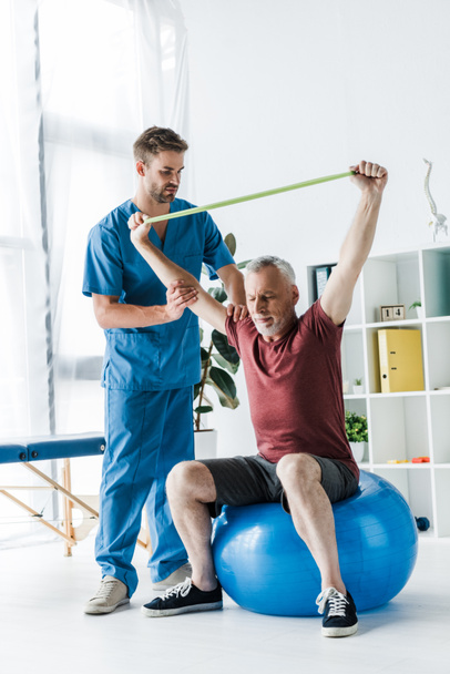 όμορφος γιατρός στέκεται κοντά μεσήλικες άνθρωπος άσκηση με μπάντα αντίστασης, ενώ κάθονται στο γυμναστήριο μπάλα  - Φωτογραφία, εικόνα