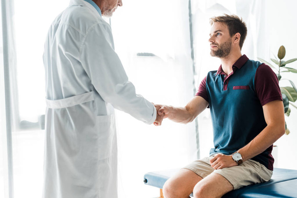 cropped view of doctor in white coat shaking hands with handsome man  - Fotoğraf, Görsel