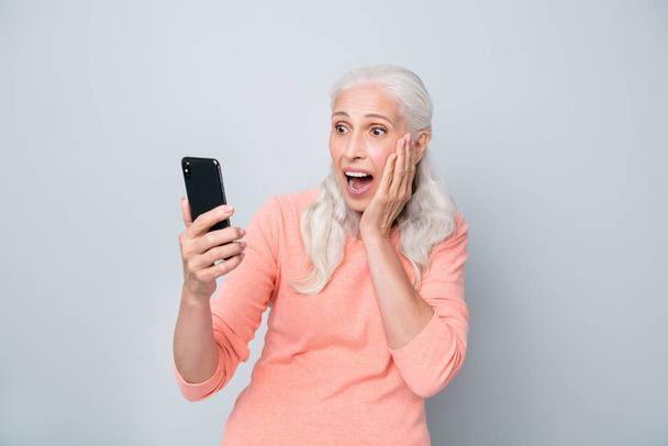 Photo of amazed charming with opened mouth old lady holding using telephone in hands isolated grey background - Foto, imagen