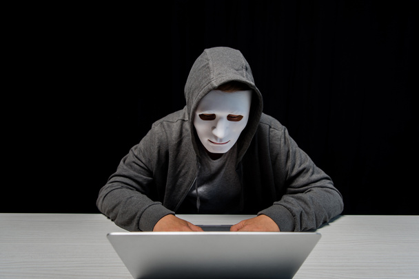 anonymous internet troll in mask typing on laptop keyboard isolated on black - Фото, изображение