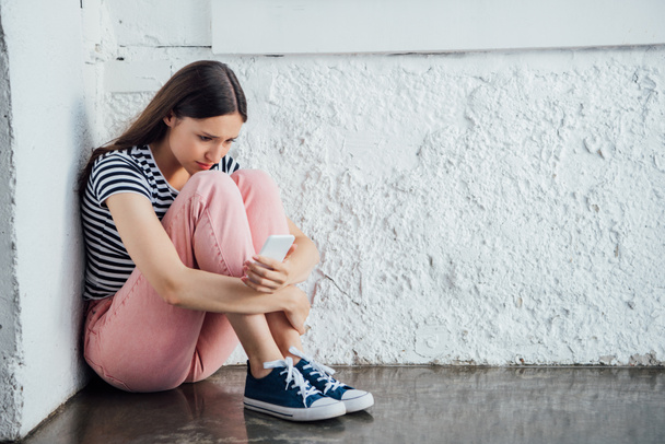 trauriges Mädchen in pinkfarbener Hose sitzt neben Wand und hält Smartphone in der Hand - Foto, Bild