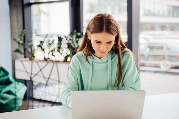 gericht meisje in Casual hoodie zittend aan tafel en met behulp van laptop - Foto, afbeelding