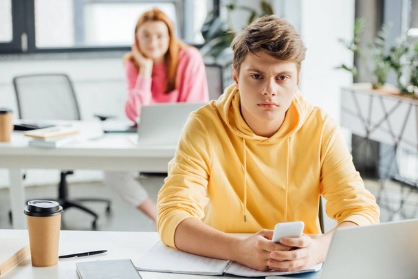 pensive boy in yellow hoodie using smartphone at desk - Photo, Image