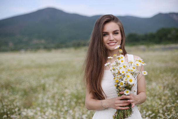 Beautiful girl outdoors with a bouquet of flowers in a field of white daisies,enjoying nature. Beautiful Model with long hair in white dress having fun on summer Field with blooming flowers,Sun Light. Young Happy Woman on spring meadow, countryside - Valokuva, kuva