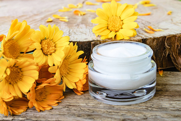 Een potje witte cosmetische crème voor lichaamsverzorging. Verse oranje Calendula bloemen op houten achtergrond. - Foto, afbeelding