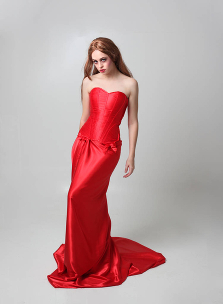 full length portrait of a  girl wearing a long red silk gown, Standing pose on a grey studio background. - Fotografie, Obrázek