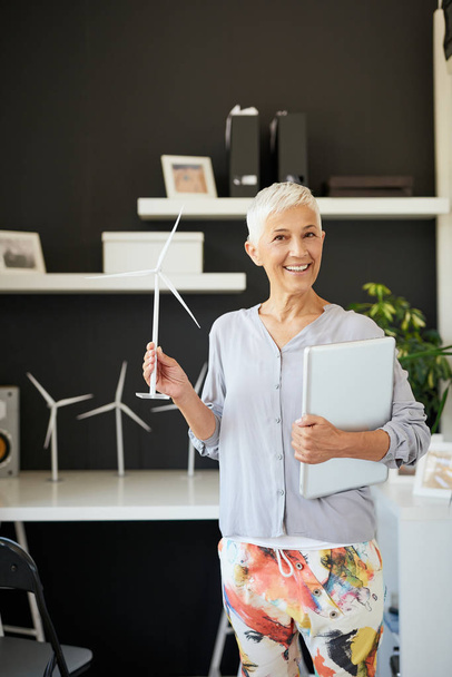 Attractive smiling Caucasian senior woman dressed casual and with short gray hair standing in office, holding laptop and windmill. Sustainable concept. - Foto, Imagen