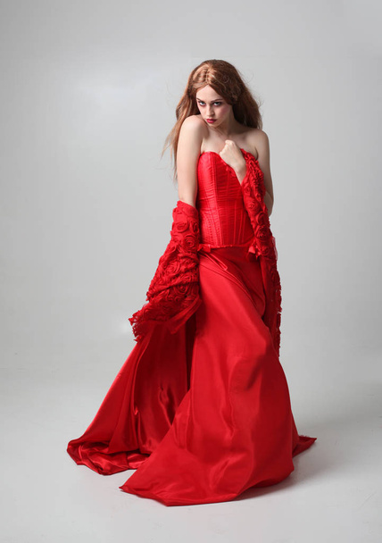 full length portrait of a  girl wearing a long red silk gown, Standing pose on a grey studio background. - Foto, afbeelding