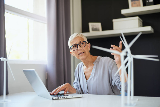 Ciddi Kafkas yaşlı kadın dizüstü bilgisayar kullanarak ve ofiste otururken meslektaşı ile konuşurken. Masa başında dizüstü bilgisayar ve yel değirmeni modelleri bulunmaktadır. Sürdürülebilir kavram. - Fotoğraf, Görsel