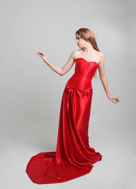 full length portrait of a  girl wearing a long red silk gown, Standing pose on a grey studio background. - Foto, afbeelding