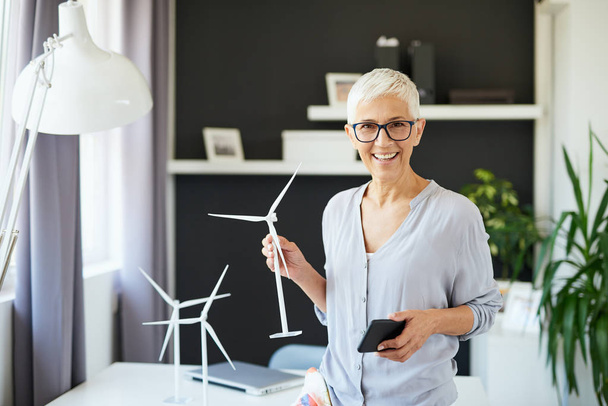 Beautiful smiling Caucasian senior woman standing in office, holding windmill model and smart phone. Start up business concept. - Fotografie, Obrázek
