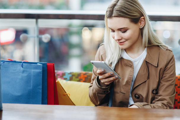 Young woman in brown cloak with shopping bags sitting in cafe at shopping mall. Girl with a phone in her hand. Buying too much concept. Happy shopping. Copy space on the left side - Foto, imagen