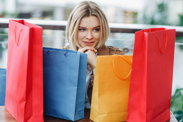 junge, zufriedene blonde Frau mit Einkaufstüten im Einkaufszentrum nach dem Einkauf. Taschen im Vordergrund. Kauf zu viel Konzept. fröhliches Einkaufen. Shopper, Verkauf, Einkaufszentrum - Foto, Bild
