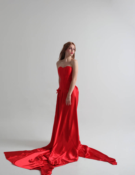 full length portrait of a  girl wearing a long red silk gown, Standing pose on a grey studio background. - Photo, image