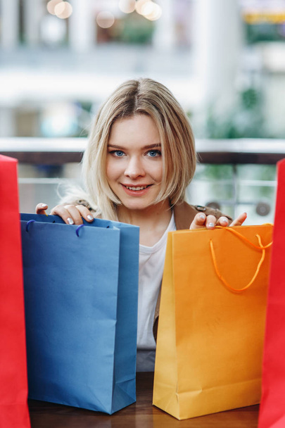 junge blonde Frau mit blauen und gelben Einkaufstaschen im Einkaufszentrum nach dem Einkaufen, in die Kamera blickend. Taschen im Vordergrund. Kauf zu viel Konzept. fröhliches Einkaufen. Kopierraum über dem Kopf - Foto, Bild
