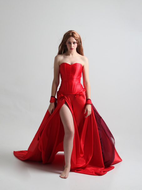 ολόσωμο πορτρέτο ενός κοριτσιού φορώντας ένα μακρύ κόκκινο μεταξωτό φόρεμα, στέκεται ποζάρει σε ένα γκρίζο φόντο στούντιο. - Φωτογραφία, εικόνα