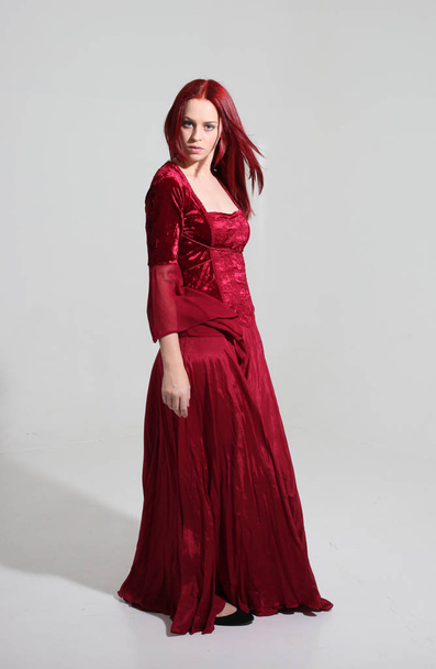 full length portrait of a  girl wearing a  red medieval fantasy gown, Standing pose on a grey studio background. - Foto, Imagem