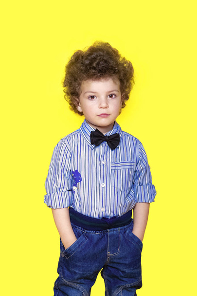 A kid portrait with curly hair on studio. Portrait of a confident smiling little kid standing with hands in pockets isolated over yellow background - Foto, Imagem