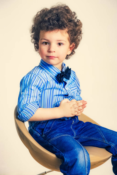 Dětský portrét se kudrnatými vlasy ve studiu. Chlapec v bílé košili, sedící na vysoké židli - Fotografie, Obrázek