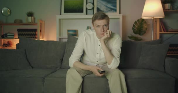Portrait of unhappy student clicking remote control watching TV in dark room - Materiał filmowy, wideo