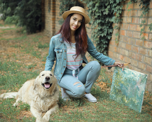 Πορτραίτο της νεαρής όμορφη 30 ετών γυναίκα καλλιτέχνης κρατώντας μια εικόνα και βούρτσες και το κατοικίδιο σκυλί της Γκόλντεν τρίτρβερ ξαπλωμένη δίπλα της στο γρασίδι υπαίθρια - Φωτογραφία, εικόνα