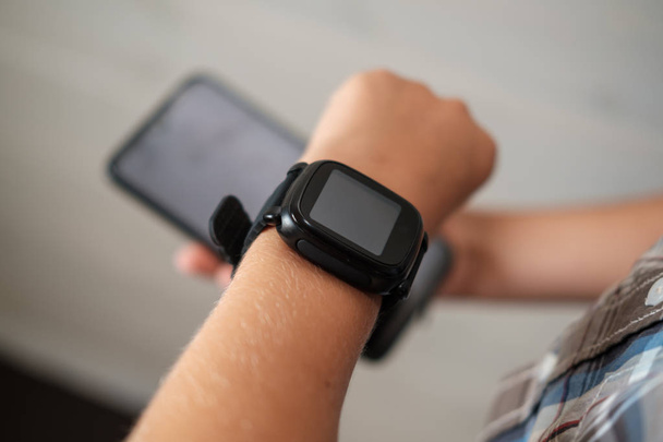 black smart watch on the boy's hand and black smartphone in the other hand - Photo, Image