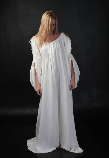 full length portrait of blonde girl wearing long white flowing robe. standing pose against a black studio background. - Foto, Imagem