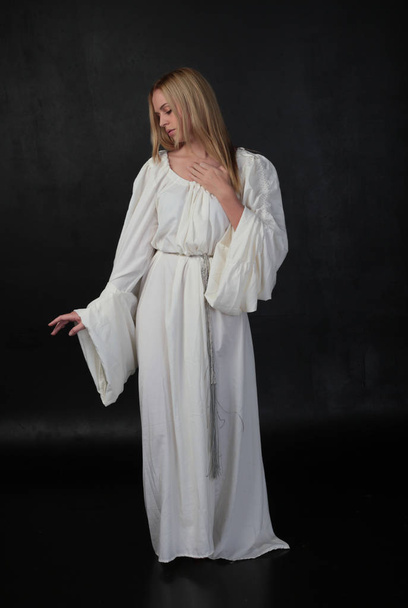 full length portrait of blonde girl wearing long white flowing robe. standing pose against a black studio background. - Photo, Image