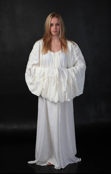 full length portrait of blonde girl wearing long white flowing robe. standing pose against a black studio background. - Фото, изображение
