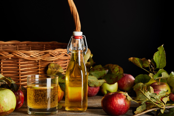 bottle and glass of homemade cider near wicker basket and apples isolated on black - Photo, Image