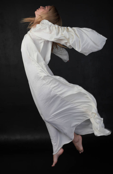 Ganzkörperporträt eines blonden Mädchens in einer langen weißen, fließenden Robe. Stehende Pose vor schwarzem Studiohintergrund. - Foto, Bild