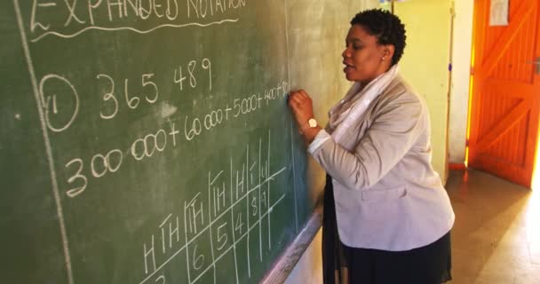 Side view close up of a middle aged African female school teacher standing at the front of the class writing on the blackboard during a maths lesson in a township elementary school classroom 4k - Video, Çekim