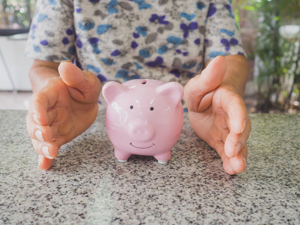 Oude vrouw hand voorkomen roze Piggy Bank staan op marmeren vloer. Geld besparen voor toekomstig plan en pensioenfonds concept - Foto, afbeelding