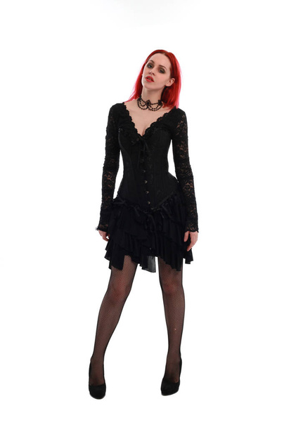 portrait complet d'une fille aux cheveux roux portant une robe gothique noire, pose debout sur un fond de studio gris
. - Photo, image