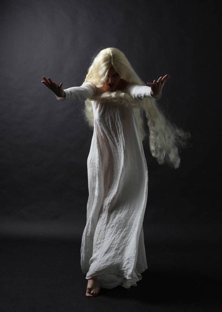Примарно повнометражний портрет жінки з довгим світлим волоссям у білому халаті. Позування на фоні чорної студії
.  - Фото, зображення