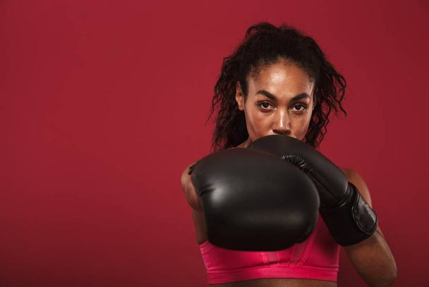 Serious strong young african sports woman boxer posing isolated over red wall background make boxing exercises. - Fotó, kép