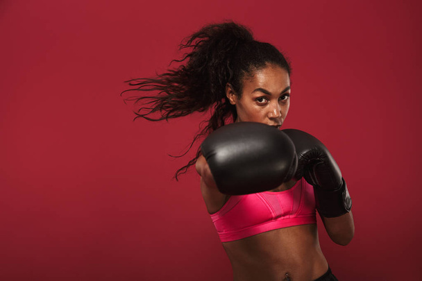 Serious strong young african sports woman boxer posing isolated over red wall background make boxing exercises. - Foto, imagen