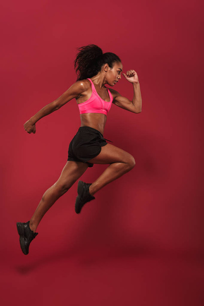 Runner concentrated strong young african sports woman posing isolated over red wall background running jumping. - Фото, зображення