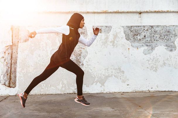 Serious muslim sports fitness woman dressed in hijab and dark clothes running outdoors. - 写真・画像