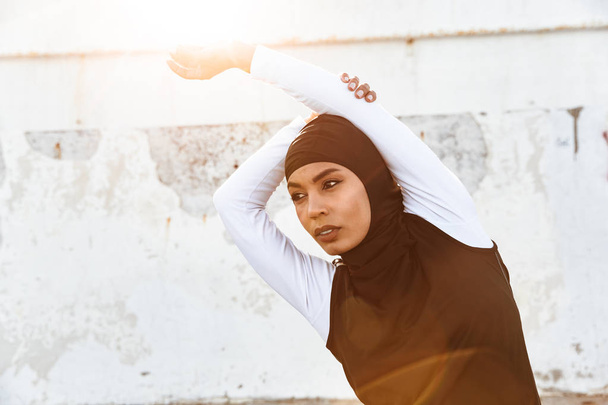 Strong muslim sports fitness woman dressed in hijab and dark clothes posing make sport stretching exercises outdoors at the street. - Fotografie, Obrázek