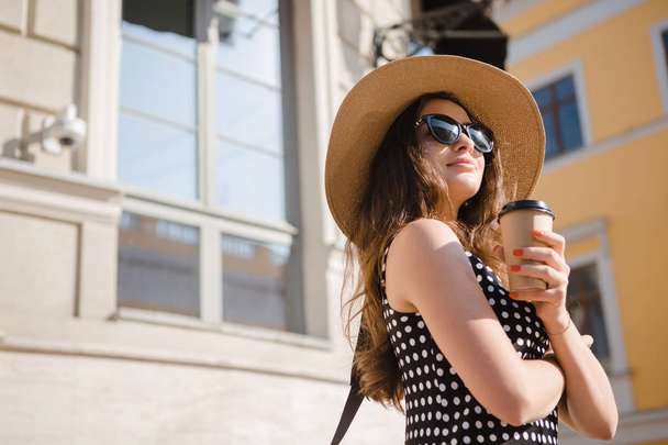 Pretty girl in a black dress, hat with wide flaps and sun glasses holds disposable paper cup of hot coffee enjoying walk down the European town during sunny summer day. - Фото, изображение