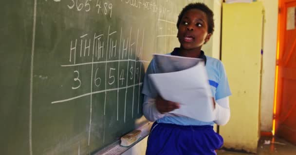 Side view close up of a young African schoolgirl standing at the front of the class reading from a book she is holding and pointing to the blackboard explaining during a lesson in a township elementary school classroom 4k - Metraje, vídeo