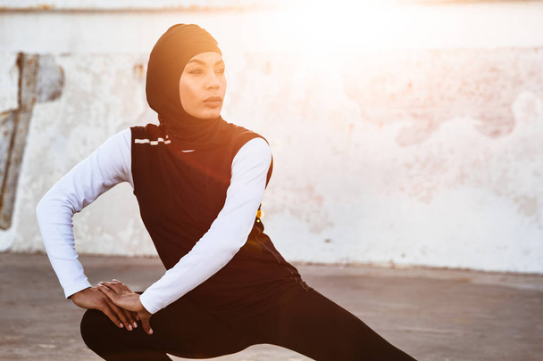 Concentrated muslim sports fitness woman dressed in hijab and dark clothes posing make sport stretching exercises outdoors at the street. - Fotó, kép