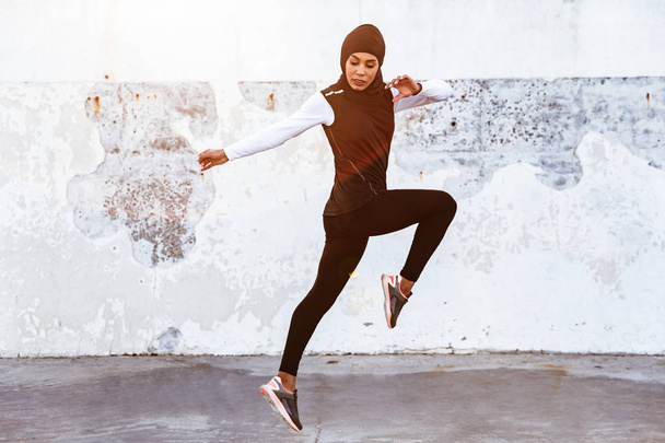 Concentrated muslim sports fitness woman dressed in hijab and dark clothes posing make sport stretching exercises outdoors at the street running. - 写真・画像