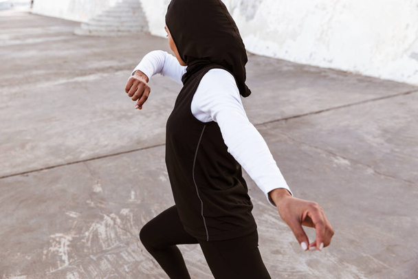 Concentrated muslim sports fitness woman dressed in hijab and dark clothes posing make sport stretching exercises outdoors at the street running. - Fotoğraf, Görsel