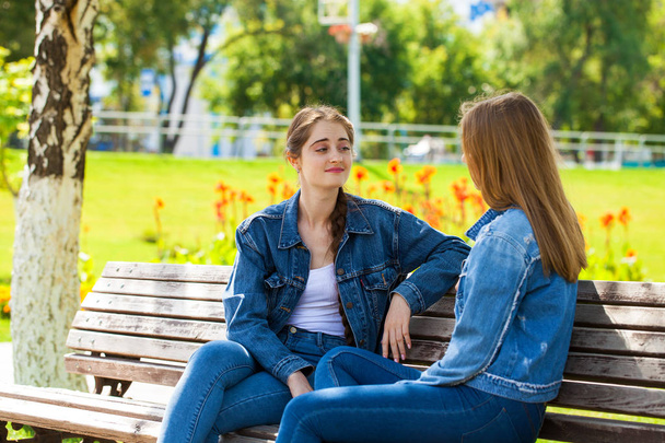 Two beautiful girlfriends are talking in a park sitting on a ben - Zdjęcie, obraz