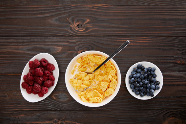 top view of bowl with cornflakes near plates with raspberry and blueberry on wooden surface - 写真・画像