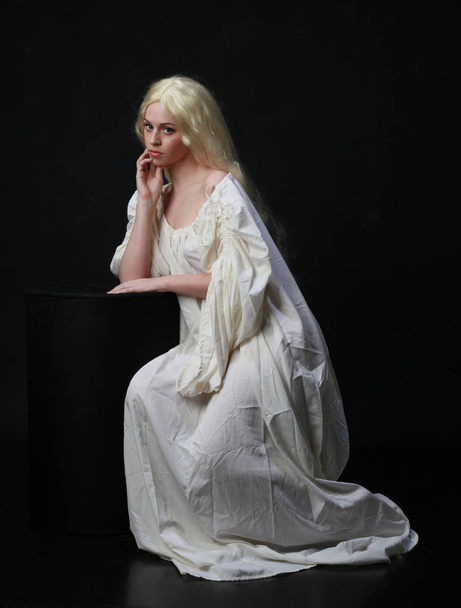 Gespenstisches Porträt einer Frau mit langen blonden Haaren, die ein weißes Gewand trägt. Posing vor schwarzem Studiohintergrund.  - Foto, Bild