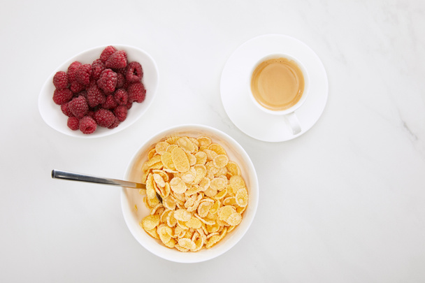 top view of bowl of cornflakes with spoon near cup of coffee and plate with raspberry on white marble surface - Fotografie, Obrázek